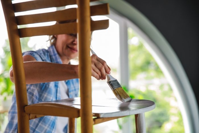 Cut out of a wooden chair that active mature woman is painting with white paint while learning a new hobbie during lockdown days at home.