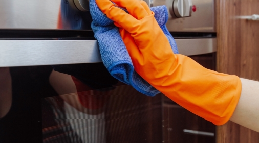 An orange-gloved hand with a microfiber cleaning cloth is being wiped on the outside of the electric oven. The cleaning of the kitchen concept