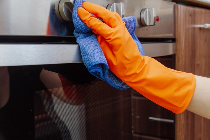 An orange-gloved hand with a microfiber cleaning cloth is being wiped on the outside of the electric oven. The cleaning of the kitchen concept